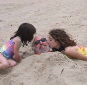 Ted at the beach with his two daughters. He is buried, except for his head. His daughters are kissing him on the cheecks.